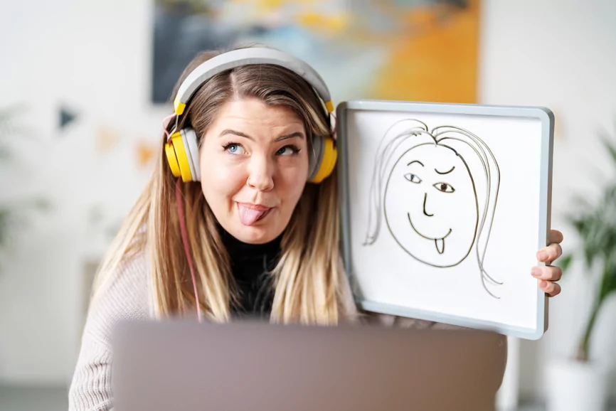 Funny female English tutor having video call on laptop and showing drawing with face expression during remote lesson